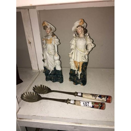 107 - A pair of glazed contiental bisque figurines and a pair of salad servers