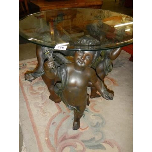 989 - A glass topped centre table supported by three cherubs. COLLECT ONLY.