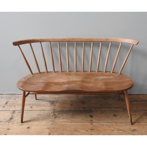 57 - AN ERCOL MID CENTURY LIGHT ELM BENCH SEAT, the gently curving top rail supported by turned tapered s... 