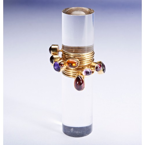 319 - WENDY RAMSHAW; AN AMETHYST, GARNET, AND GOLD RING ON AN ACRYLIC STANDthe ring composed of twelve ban... 