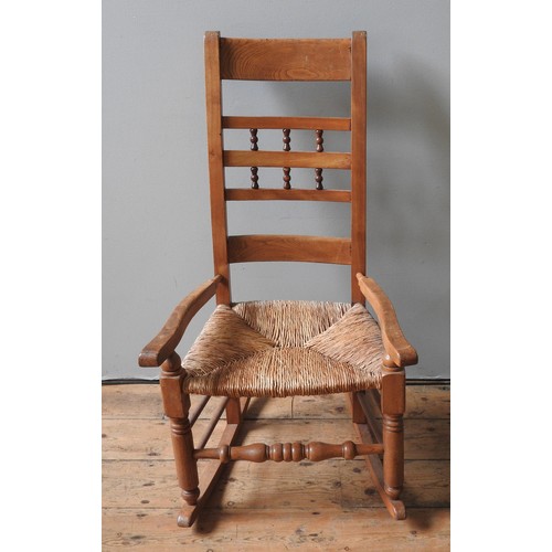 33 - A PINE LADDER BACK ROCKING CHAIR, the rush seat panel flanked by low arms and raised on turned woode... 