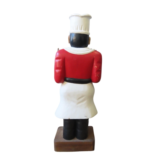 454 - A PAINTED HARDWOOD FIGURE OF A CHEF, modelled standing, holding a spoon and fork, raised on a rectan... 