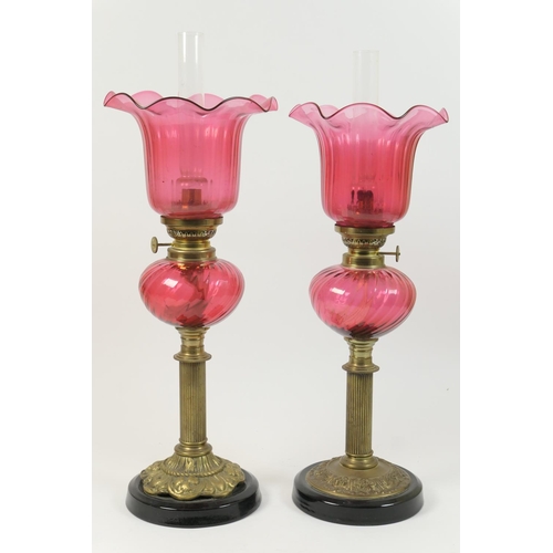 443 - Two similar cranberry glass and brass pedestal oil lamps, each having a moulded bell shaped shade ov... 