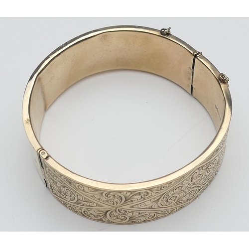 409 - 9ct gold hinged bangle, chased with foliate scrolls, with safety chain, interior size approx. 54mm, ... 