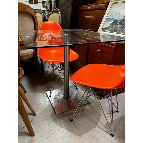 987 - A square glass kitchen table with brushed steel pedestal, two bright orange dining chairs 