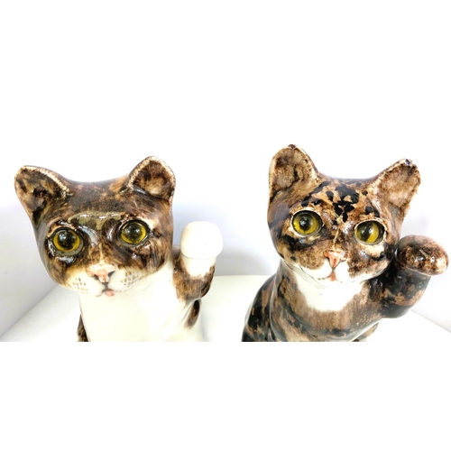 561 - Two Winstanley cats, glass eyes, both seated with one paw raised,  signed, size 3 (each a.f)