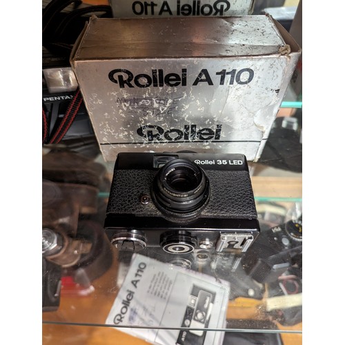 730 - Three shelves of cameras and accessories inc. Rollei 35 LED, Rollei A110, Konica, Pentax, Samsung et... 