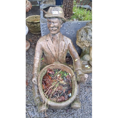 87 - A figural cast stone garden planter modelled as a seated gentleman in hat and suit, holding a circul... 