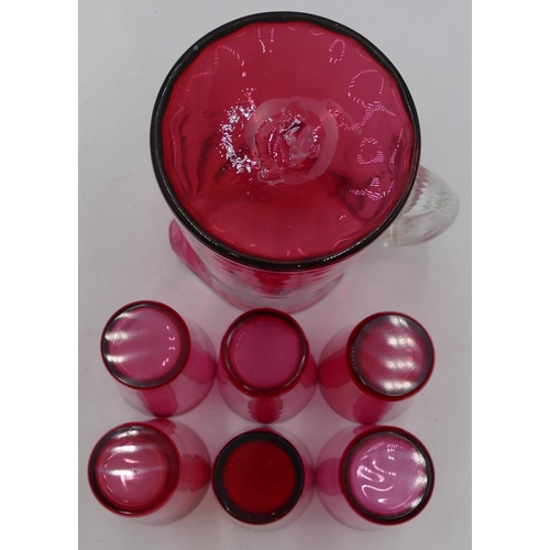 64 - A Cranberry glass round jug with clear reeded handle, 18cm high and 6 Cranberry beakers. (7)