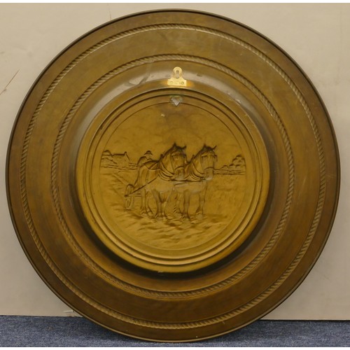 7009 - A large brass charger with embossed scene of horses ploughing to the centre and hanging fixing to ba... 