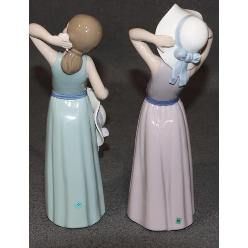 133 - 2 Lladro figures of standing young ladies holding hats wearing purple and green dresses, largest 26c... 