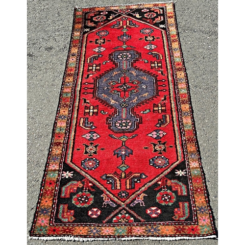 1584 - A narrow Kazak runner with a central blue extended diamond medallion and stylised flowers on a red g... 