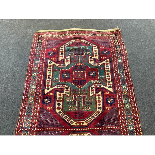 196 - An antique Kazak red ground rug, with double shield motif within a hooked geometric field and triple... 