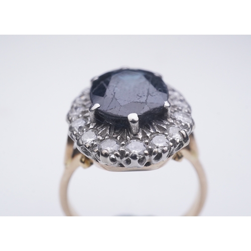 367 - A sapphire and diamond ring, of cluster design, claw-set with an oval deep blue/green sapphire weigh... 