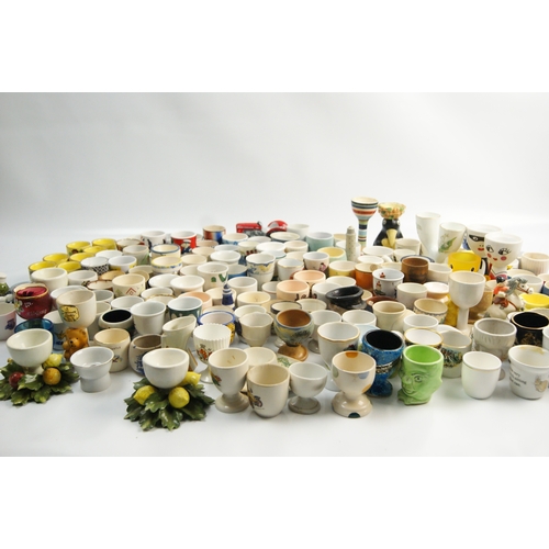 65 - A Large Collection of Ceramic and Glass Egg Cups Including Novelty and Souvenir ExamplesAn extensi... 