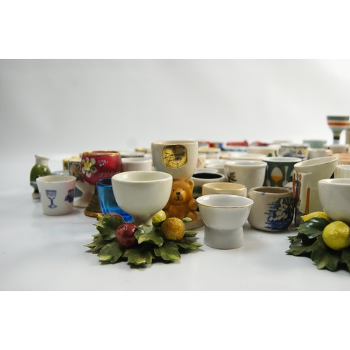 65 - A Large Collection of Ceramic and Glass Egg Cups Including Novelty and Souvenir ExamplesAn extensi... 