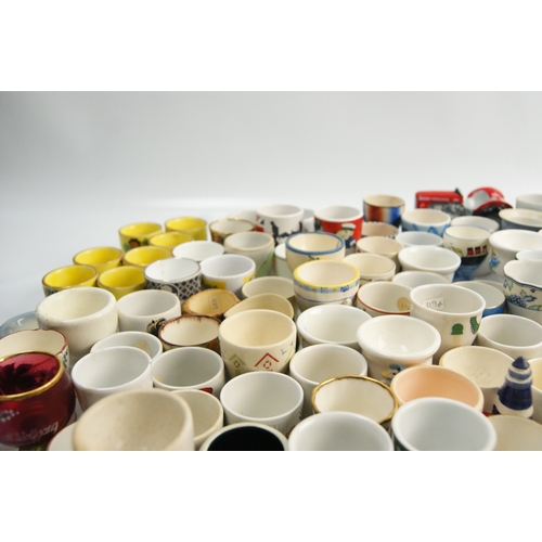65 - A Large Collection of Ceramic and Glass Egg Cups Including Novelty and Souvenir ExamplesAn extensi... 