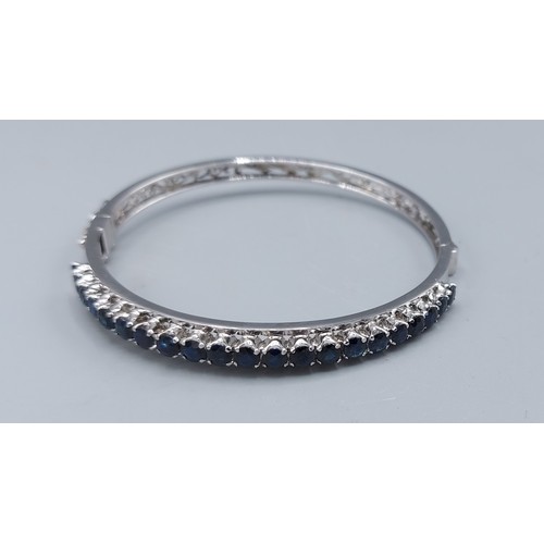 211 - A white metal bangle set with a row of twenty one sapphire, claw set, 12.9gms, 6.5cms diameter