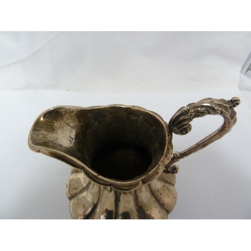 16 - A silver jug, the body of lobed squat baluster form with high foliate scroll handle and upon four vo... 