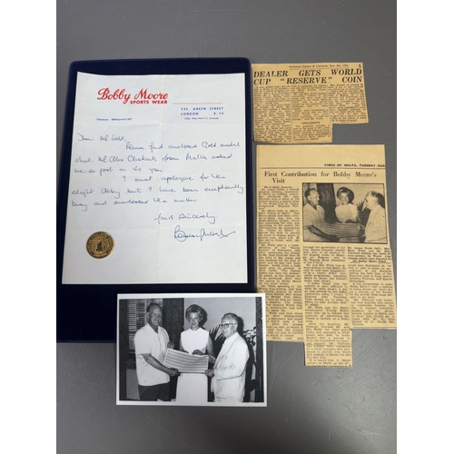 Bobby Moore Personal Hand-Signed Letter and Rare 1966 Maltese Gold Commemorative Medal Provenance Archive. 14.96grams.

A significant historical memorabilia archive documenting the connection between England’s 1966 World Cup-winning captain, Bobby Moore, and the island of Malta. This lot includes a personal handwritten letter, a rare gold medal, and extensive contemporary supporting documentation providing exceptional provenance.

The personal handwritten letter is on Bobby Moore Sports Wear company letterhead, addressed from 542 Green Street, London E.13, situated opposite the West Ham F.C. Ground. The letter is addressed to Mr. Webb and states: Please find enclosed Gold medal which Mr. Alex Chetcuti from Malta asked me to post on to you. I must apologise for the slight delay but I have been exceptionally busy and overlooked the matter. The letter is hand-signed in blue ink, Bobby Moore.

The accompanying gold commemorative medal was minted for the 400th anniversary of the founding of Valletta (1566–1966). The obverse features a profile portrait of Grand Master Jean Parisot de Valette with a Maltese cross and the inscription CONSOCIATIO MELITENSIS S.M.O.M. The reverse displays fortifications with the text CCCC AN. A VALLETTA CONDITA and DEO JUV, dated 1566-1966. The medal is marked 900/1000 on the reverse, indicating 21.6 carat gold purity, and bears the maker mark ARGOR - NU for Argor SA, Switzerland. According to contemporary press accounts included in the lot, this medal is one of a limited issue of only twelve minted.

Supporting ephemera includes an original black and white press photograph showing Mr. James Webb, Mrs. Ruby Webb, and Mr. Alex Chetcuti holding a sheet of 120 England Winners overprinted World Cup stamps. The archive is completed by several contemporary newspaper clippings, including the Doncaster Gazette & Chronicle (8 September 1966) titled Dealer Gets World Cup Reserve Coin, and the Times of Malta (August 1966) titled First Contribution for Bobby Moore's Visit. A translation of the article in the Maltese language is also present. Memorabilia

Weight: 14.96g.