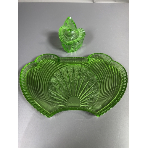 A Pair of Samuel Reich Viktoria Pattern Uranium Glass Vanity Items

A two-piece dressing table or vanity set consisting of a crescent-shaped tray and a matching lidded heart-shaped trinket box. Both pieces are manufactured from translucent green uranium glass, which fluoresces under ultraviolet light. The set features the Viktoria pattern, characterised by a central radiating fan or sunburst motif set against a stippled hobnail ground. The tray is designed with wide wings featuring concentric arced ridges and a vertically fluted rim. The heart-shaped box has vertically fluted sides and a matching lid topped with a heart-shaped finial decorated with a sunburst design. This set dates to the 1930s Art Deco period and is a product of the Samuel Reich glassworks. No maker marks or signatures are present.
