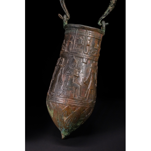 An Egyptian Bronze Funerary Vessel with Anubis and Royal Iconography, Late Period, 26th Dynasty (c. 664–525 B.C.).

A rare and finely cast Egyptian bronze vessel with a twisted loop handle, possibly used for ritual libations or funerary offerings. The tapering ovoid body is richly adorned in raised relief with a ceremonial scene featuring a seated pharaoh or high-ranking figure, approached by the jackal-headed god Anubis. Anubis is shown gently holding the hand of a standing priest, an evocative representation of psychopompic guidance—leading the soul into the afterlife. Additional registers include figures in adoration and hieratic poses, all framed within stylised architectural borders. The vessel’s lower section is decorated with incised geometric motifs, converging to a pointed finial. Likely intended for a high-status burial.

H: Approximately 40.5cm
H (Without handle): Approximately 27cm

Provenance: Part of a private collection based in W London.