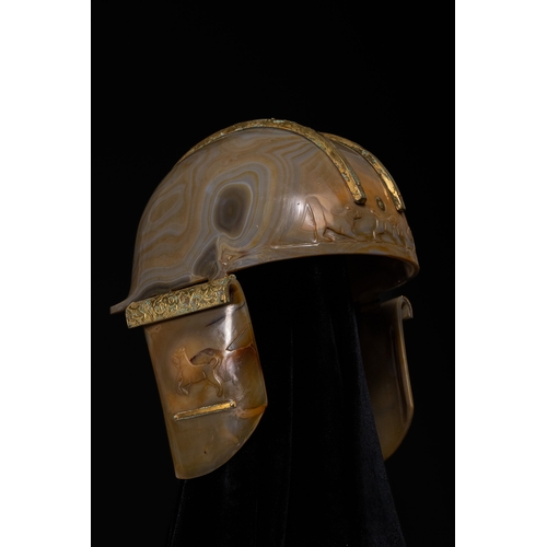 A Rare Chinese Agate Helmet with Gold Gilt-Metal Mounts and Figural Friezes, 18th Century.

A helmet-form object carved from banded agate, the domed body displaying natural swirling layers of grey, honey, and amber tones. The surface is carved in low relief with a continuous frieze of running animals encircling the lower register. The sides are fitted with hinged cheek guards, each similarly carved with a stylised animal in relief. Gilt-metal mounts, including twin arched bands across the crown and decorative fittings to the sides, provide structural and visual contrast.

H: Approximately 16cm