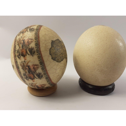 268 - Two OSTRICH EGGS on stands one decorated with maps of Africa and African WILDLIFE and the other unde... 