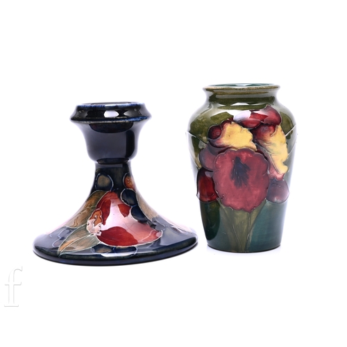 William Moorcroft for Moorcroft Pottery, a Pomegranate pattern candlestick, tube line decorated with fruit and berries in tonally red and green over blue ground, impressed mark and facsimile signature to base, height 8cm, and Orchid pattern vase, tube line flower heads in tonal lemon and red, over green ground, printed label to base, height 9.5cm. (2)