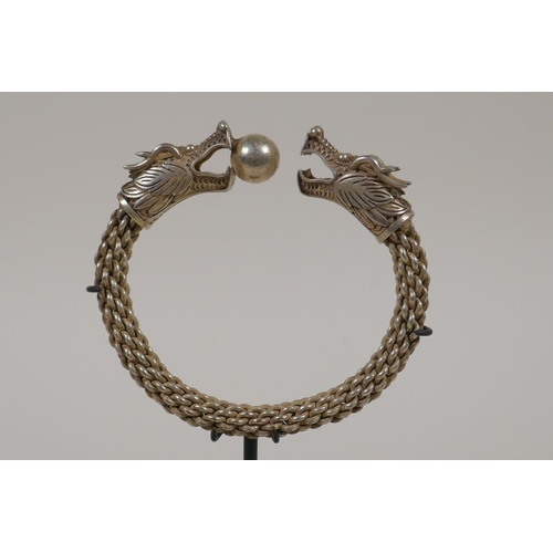 28 - A Chinese white metal rope twist bangle with twin dragon head decoration to ends, on a display stand... 