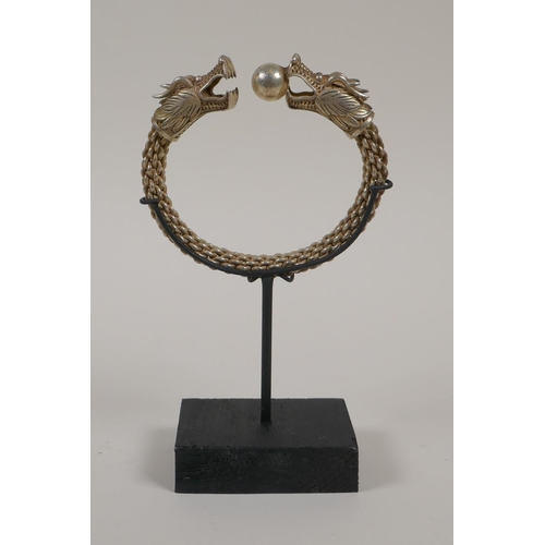 28 - A Chinese white metal rope twist bangle with twin dragon head decoration to ends, on a display stand... 