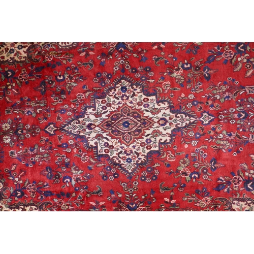 1099 - A Persian full pile red ground hand woven Sarouk village carpet with a floral medallion pattern and ... 