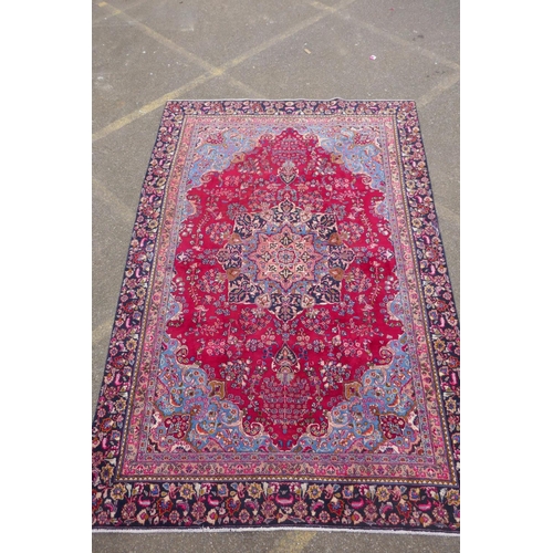 1065 - A claret and blue ground Persian Mashad carpet with a traditional floral medallion design, patched, ... 