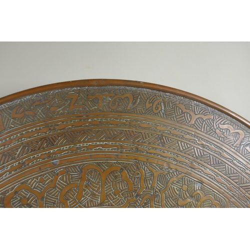 1172 - A Persian brass tray table on a folding base, with engraved Islamic script decoration, 60cm diameter... 