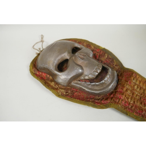 178 - A Tibetan white metal citipati death mask on a silk mount, 45cm long