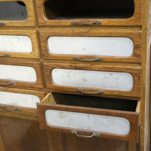 3127 - An early 20th century oak shopfitting haberdashery cabinet, having sixteen drawers above a cupboard ... 