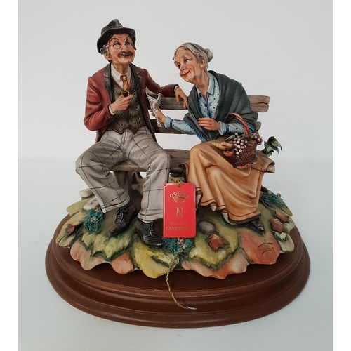 CAPODIMONTE FIGURINE
depicting an old couple sat on a park bench, the gentleman smoking his pipe, the lady with a basket of fruit, on an oval mahogany plinth, 26cm high