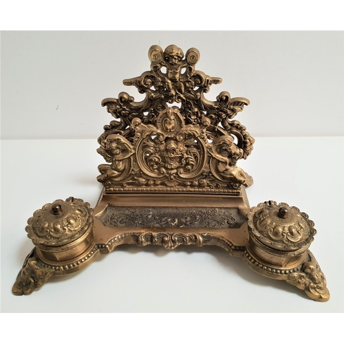 191 - ORNATE BRASS DESK STAND
with a rear letter rack decorated with putti, with two circular lidded inkwe... 