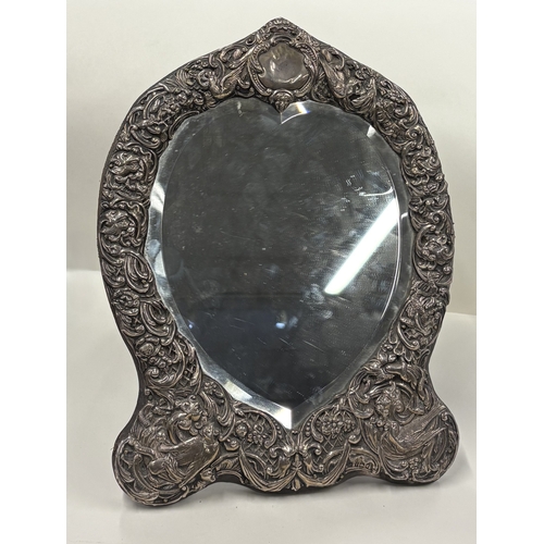 700 - A large heart-shaped silver-mounted mirror profusely decorated with birds and scrolls, with vacant c... 