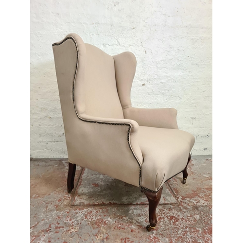 412 - An early 20th century beige upholstered wingback armchair with mahogany supports and casters - appro... 