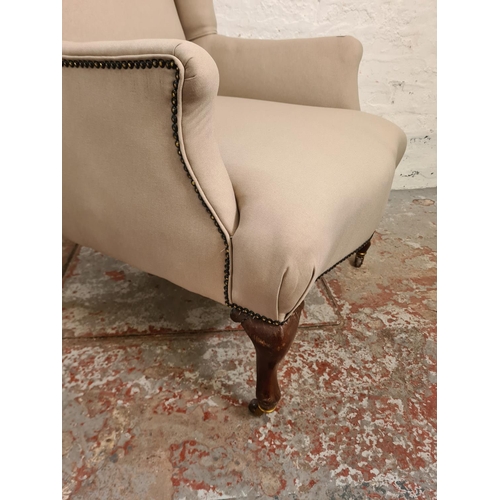 412 - An early 20th century beige upholstered wingback armchair with mahogany supports and casters - appro... 