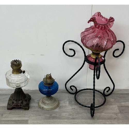 128 - Three oil lamps, one cranberry glass and brass on wrought metal stand, one British made metal and bl... 