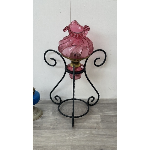 128 - Three oil lamps, one cranberry glass and brass on wrought metal stand, one British made metal and bl... 