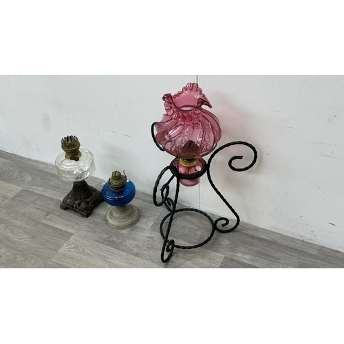 128 - Three oil lamps, one cranberry glass and brass on wrought metal stand, one British made metal and bl... 