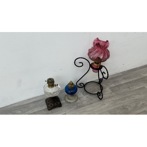 128 - Three oil lamps, one cranberry glass and brass on wrought metal stand, one British made metal and bl... 