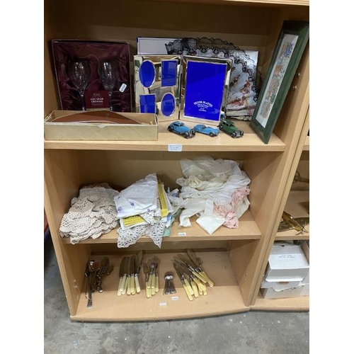 3 shelves of collectables inc. mixed cutlery, linens & lace, 3 Dinky Toys, 2 boxed cut lead crystal sherry glasses, boxed Harp lager boomerang clothes brush etc.