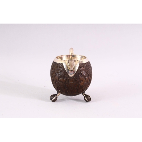 841 - A FINELY CARVED THAI COCONUT JUG WITH SILVER MOUNTS, on three ball feet, 10cm high .