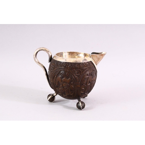 841 - A FINELY CARVED THAI COCONUT JUG WITH SILVER MOUNTS, on three ball feet, 10cm high .