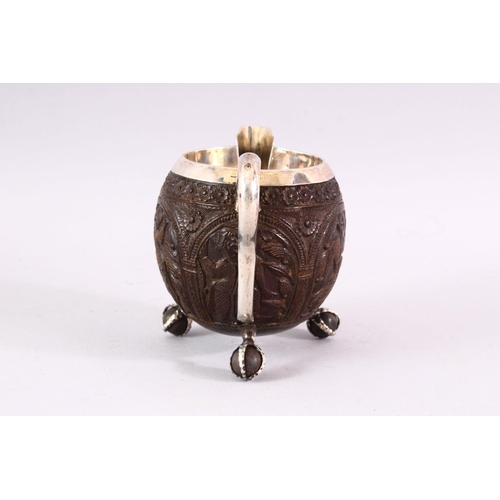 841 - A FINELY CARVED THAI COCONUT JUG WITH SILVER MOUNTS, on three ball feet, 10cm high .