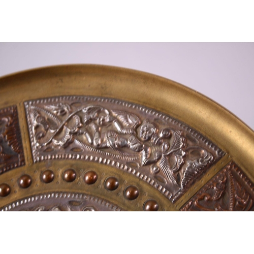 613 - AN INDIAN BRASS CIRCULAR DISH, overlaid with white metal and copper panels, the centre depicting a f... 